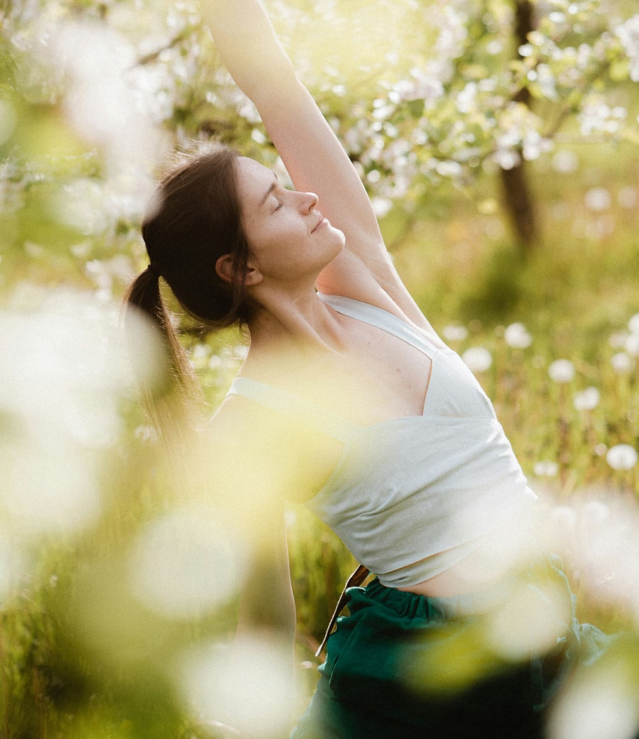 Woman stretching outdoors in a field of blooming flowers, surrounded by soft sunlight and blurred blossoms, wearing a light grey top and dark green pants.