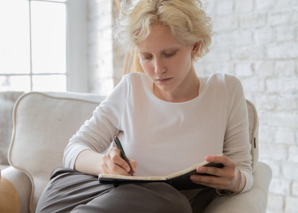 Woman journaling on a cosy armchair in a light-filled living room as part of a stress relief routine for mental wellness